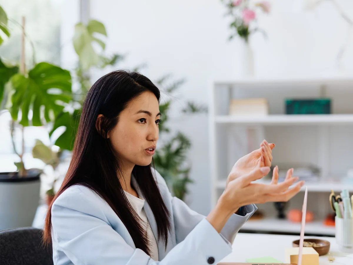 Nữ quản lý người châu Á tóc dài, mặc blazer xanh nhạt, đang giải thích ý tưởng bằng cử chỉ tay trong văn phòng sáng có cây xanh.